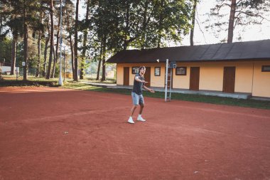 Yüzünde gülümseme olan siyah saçlı bir sporcu kil sahada badminton oynar. Amatör spora olan tutkum. Yeni hareket becerileri öğreniliyor.