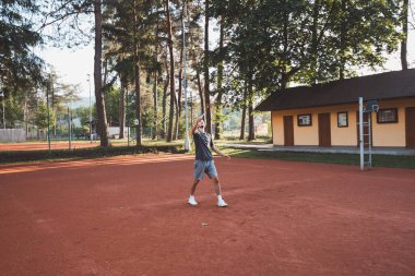 Yüzünde gülümseme olan siyah saçlı bir sporcu kil sahada badminton oynar. Amatör spora olan tutkum. Yeni hareket becerileri öğreniliyor.