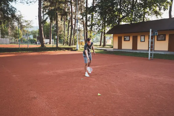 Yüzünde gülümseme olan siyah saçlı bir sporcu kil sahada badminton oynar. Amatör spora olan tutkum. Yeni hareket becerileri öğreniliyor.