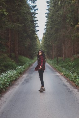 Kırmızı ekose gömlekli genç bir kadın ağaçlarla kaplı, huzurlu bir yolda yürüyor, doğanın sükunetini ve güzelliğini kucaklıyor, özgürlüğü ve kendini keşfetme yolculuğunu simgeliyor..