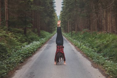 Acrobatic woman in black leggings and plaid shirt performs a cartwheel on a forest path in the middle of a deep forest. Coordination and agility training. clipart