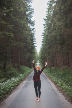Kırmızı kareli gömlekli, parlak turuncu bereli gülümseyen kadın orman yolunun ortasında duruyor, kollarını başının arkasına uzatıyor, çevrenin sakinliğinin tadını çıkarıyor..