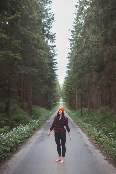 Kırmızı kareli gömlekli, parlak turuncu bereli gülümseyen kadın orman yolunun ortasında duruyor, kollarını başının arkasına uzatıyor, çevrenin sakinliğinin tadını çıkarıyor..
