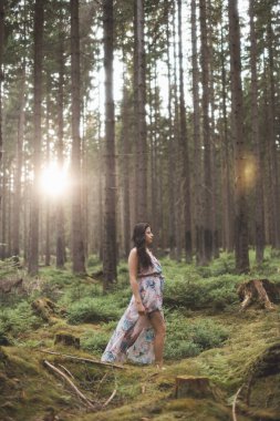 20-25 yaşlarındaki güzel esmer kız renkli bir elbiseyle ormanda dans ediyor. Temiz havanın neşesi ve doğanın gücü. Orman Perisi.