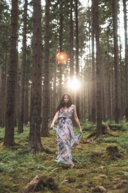 20-25 yaşlarındaki güzel esmer kız renkli bir elbiseyle ormanda dans ediyor. Temiz havanın neşesi ve doğanın gücü. Orman Perisi.