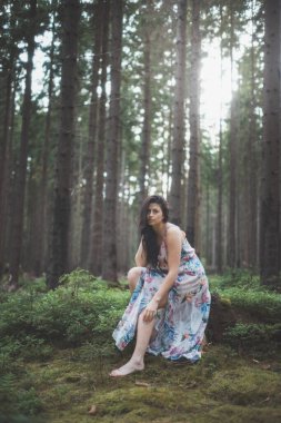 20-25 yaşlarındaki güzel esmer kız renkli bir elbiseyle ormanda oturuyor. Temiz havanın neşesi ve doğanın gücü. Orman perisi. Güzel ve şefkatli hissediyorum.