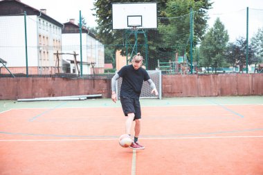 Güneşli bir günde, turuncu sahada topuyla hokkabazlık yapan genç bir adam. Spor kıyafetleri giyerek, topu doğal bir ortamda hassas bir şekilde kontrol etmeye odaklanıyor.