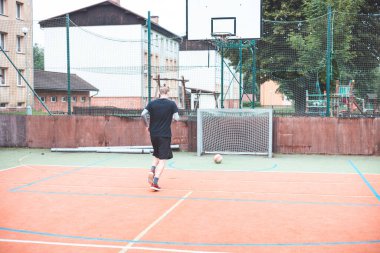 Genç adam kaleye doğru koşuyor, bir futbol topuna hazırlanıyor. Açık hava sahasında binalar ve basketbol potası olan bir kent zeminine, gündelik sporlarla odak ve enerjiyi birleştiren bir basketbol potası yer alıyor..