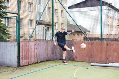 Genç bir adam açık havada top havadayken güçlü bir futbol tekmesi atıyor. Günlük atletizm kıyafetleri giyiyor, şehir spor ortamında odaklanma ve enerji gösteriyor..
