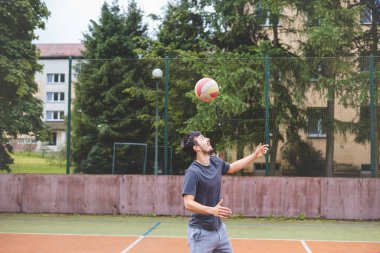 Genç adam açık havada bir sahada futbol topuna yönelirken odaklanıyor. Günlük spor kıyafetleri giyerek, arka planda ağaçlar olan hareketli bir sahnede denge ve teknik sergiliyor..