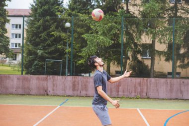 Genç adam açık havada bir sahada futbol topuna yönelirken odaklanıyor. Günlük spor kıyafetleri giyerek, arka planda ağaçlar olan hareketli bir sahnede denge ve teknik sergiliyor..