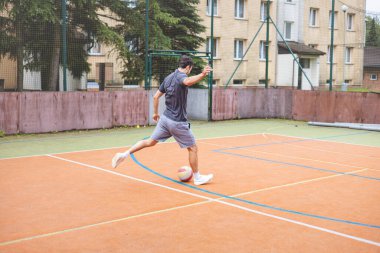 Genç adam dışarıda futbol oynarken kaleye güçlü bir atış yapıyor. Günlük giysiler içinde, yoğun bir yoğunluk gösteriyor ve arkasında kentsel bir zemin olan dinamik bir ana odaklanıyor..