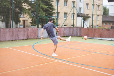 Genç adam dışarıda futbol oynarken kaleye güçlü bir atış yapıyor. Günlük giysiler içinde, yoğun bir yoğunluk gösteriyor ve arkasında kentsel bir zemin olan dinamik bir ana odaklanıyor..