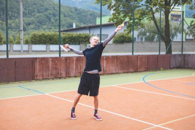 Badminton oyuncusu elinde raket ve mekikle açık bir sahada hizmet etmeye hazır. Spor kıyafetleri içinde, doğal manzaralı bir ortamda maça odaklanıyor..