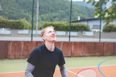 Bir badminton oyuncusunun konsantrasyonuna bakarken yakın plan görüntüsü. Maç sırasında mekiklerin horozunu takip ediyor. İfadesi, doğal ve kentsel çevre tarafından çerçevelenmiş odaklanma ve kararlılık gösteriyor..