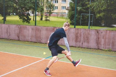 Badminton oyuncusu açık sahada çevik, düşük geri dönüş atışı yapıyor, esneklik ve hızlı refleksler gösteriyor. Spor kıyafetlerini giyip, mekiklere dikkatle vurmaya odaklanmış..