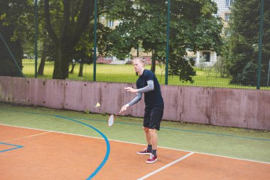 Badminton oyuncusu, açık hava sahasında oynanan bir maçta spiker horozuna vuruyor. Açık alanda kesin teknik ve atletik hareketler sergiliyor. Oyuncu ağaçlar ve kentsel bir zemin ile çevrili..