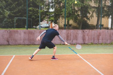 Badminton oyuncusu, açık hava sahasında oynanan bir maçta spiker horozuna vuruyor. Açık alanda kesin teknik ve atletik hareketler sergiliyor. Oyuncu ağaçlar ve kentsel bir zemin ile çevrili..