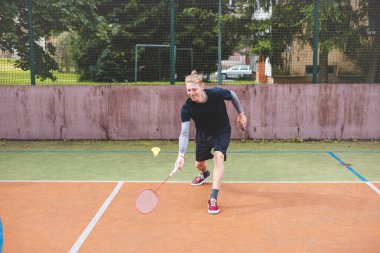 Badminton oyuncusu, açık hava sahasında oynanan bir maçta spiker horozuna vuruyor. Açık alanda kesin teknik ve atletik hareketler sergiliyor. Oyuncu ağaçlar ve kentsel bir zemin ile çevrili..