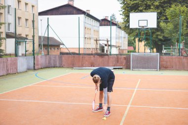 Badminton oyuncusu açık hava sahasındaki bir maçta pipisi kaldırıyor. Gündelik an, çevre binalar ve arka planda basketbol potası olan şehir ortamında geçiyor..