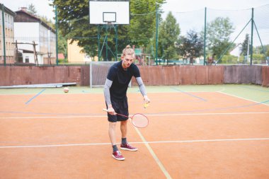 Badminton oyuncusu elinde raket ve mekikle açık bir sahada hizmet etmeye hazır. Spor kıyafetleri içinde, doğal manzaralı bir ortamda maça odaklanıyor..