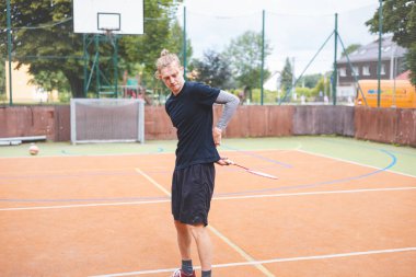 Badminton player strikes the shuttlecock during a game on an outdoor court, showcasing precise technique and athletic movement. The player is focused, surrounded by trees and an urban backdrop.