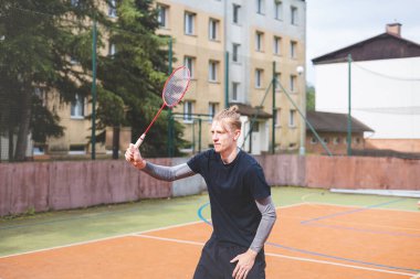 Badminton oyuncusu açık hava sahasında oynanan bir maçta havadaki pipiye odaklanıyor. Raketi yükselirken, kentsel ve manzaralı bir arka planla çevrili bir sonraki çekim için hazırlanıyor..