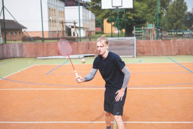 Badminton player strikes the shuttlecock during a game on an outdoor court, showcasing precise technique and athletic movement. The player is focused, surrounded by trees and an urban backdrop.