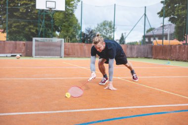 Badminton oyuncusu, açık hava sahasında oynanan yoğun bir maçta küçük bir top almak için geriliyor. Atletik duruşu odak ve kararlılık gösteriyor, arka planda kentsel bir ortam var..