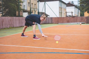 Badminton oyuncusu, açık hava sahasında oynanan yoğun bir maçta küçük bir top almak için geriliyor. Atletik duruşu odak ve kararlılık gösteriyor, arka planda kentsel bir ortam var..