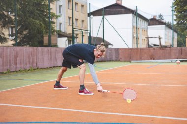 Badminton oyuncusu, açık hava sahasında oynanan yoğun bir maçta küçük bir top almak için geriliyor. Atletik duruşu odak ve kararlılık gösteriyor, arka planda kentsel bir ortam var..
