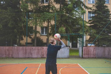 Basketbolcu topu dışarıda bir sahada atmaya hazırlanıyor ve kendini odaklanmış bir atış için hazırlıyor. Sahne, ağaçların ve kentsel binaların arka planıyla atleti hareketin ortasında yakalıyor..