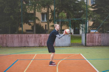 Basketbolcu topu dışarıda bir sahada atmaya hazırlanıyor ve kendini odaklanmış bir atış için hazırlıyor. Sahne, ağaçların ve kentsel binaların arka planıyla atleti hareketin ortasında yakalıyor..