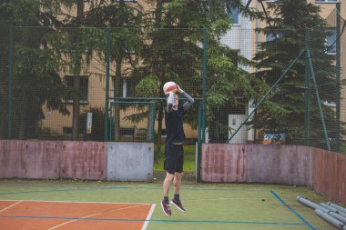 Basketbolcu topu dışarıda bir sahada atmaya hazırlanıyor ve kendini odaklanmış bir atış için hazırlıyor. Sahne, ağaçların ve kentsel binaların arka planıyla atleti hareketin ortasında yakalıyor..
