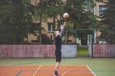 Basketbolcu topu dışarıda bir sahada atmaya hazırlanıyor ve kendini odaklanmış bir atış için hazırlıyor. Sahne, ağaçların ve kentsel binaların arka planıyla atleti hareketin ortasında yakalıyor..