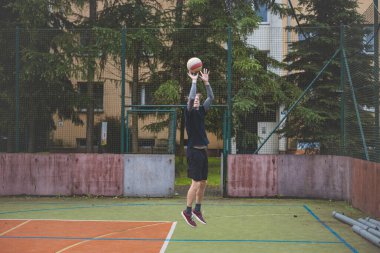 Basketbolcu topu dışarıda bir sahada atmaya hazırlanıyor ve kendini odaklanmış bir atış için hazırlıyor. Sahne, ağaçların ve kentsel binaların arka planıyla atleti hareketin ortasında yakalıyor..