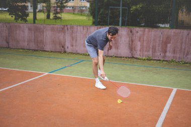 Badminton oyuncusu, açık hava sahasında oynanan yoğun bir maçta küçük bir top almak için geriliyor. Atletik duruşu odak ve kararlılık gösteriyor, arka planda kentsel bir ortam var..
