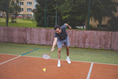 Badminton oyuncusu, açık hava sahasında oynanan yoğun bir maçta küçük bir top almak için geriliyor. Atletik duruşu odak ve kararlılık gösteriyor, arka planda kentsel bir ortam var..