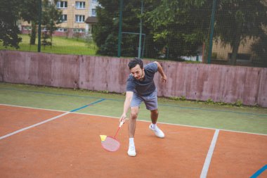 Badminton oyuncusu, açık hava sahasında oynanan yoğun bir maçta küçük bir top almak için geriliyor. Atletik duruşu odak ve kararlılık gösteriyor, arka planda kentsel bir ortam var..