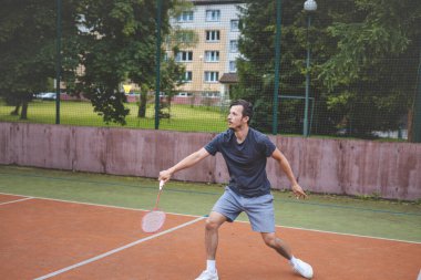 Badminton oyuncusu açık hava sahasında oynanan bir maçta havadaki pipiye odaklanıyor. Raketi yükselirken, kentsel ve manzaralı bir arka planla çevrili bir sonraki çekim için hazırlanıyor..