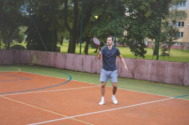 Badminton oyuncusu açık hava sahasında oynanan bir maçta havadaki pipiye odaklanıyor. Raketi yükselirken, kentsel ve manzaralı bir arka planla çevrili bir sonraki çekim için hazırlanıyor..