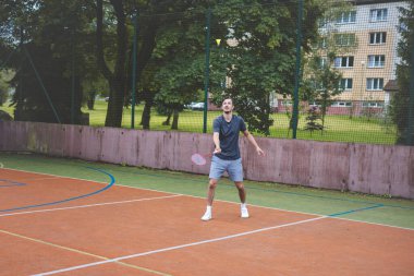 Badminton oyuncusu açık hava sahasında oynanan bir maçta havadaki pipiye odaklanıyor. Raketi yükselirken, kentsel ve manzaralı bir arka planla çevrili bir sonraki çekim için hazırlanıyor..