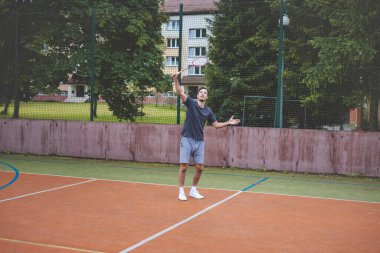 Badminton oyuncusu açık hava sahasında oynanan bir maçta havadaki pipiye odaklanıyor. Raketi yükselirken, kentsel ve manzaralı bir arka planla çevrili bir sonraki çekim için hazırlanıyor..