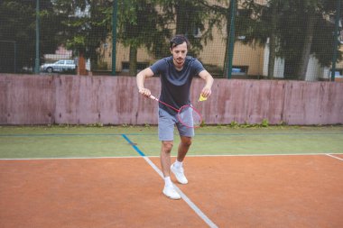 Badminton oyuncusu elinde raket ve mekikle açık bir sahada hizmet etmeye hazır. Spor kıyafetleri içinde, doğal manzaralı bir ortamda maça odaklanıyor..