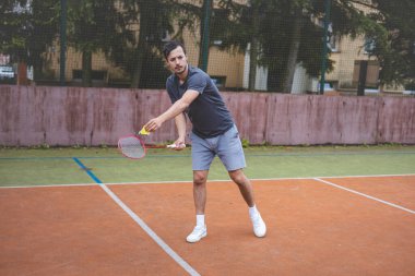 Badminton oyuncusu elinde raket ve mekikle açık bir sahada hizmet etmeye hazır. Spor kıyafetleri içinde, doğal manzaralı bir ortamda maça odaklanıyor..