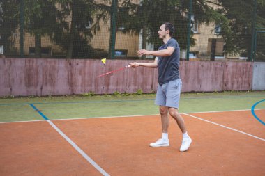 Badminton oyuncusu açık hava sahasında oynanan bir maçta havadaki pipiye odaklanıyor. Raketi yükselirken, kentsel ve manzaralı bir arka planla çevrili bir sonraki çekim için hazırlanıyor..