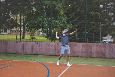 Badminton oyuncusu, açık hava sahasında oynanan bir maçta spiker horozuna vuruyor. Açık alanda kesin teknik ve atletik hareketler sergiliyor. Oyuncu ağaçlar ve kentsel bir zemin ile çevrili..