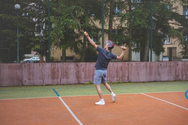 Badminton oyuncusu, açık hava sahasında oynanan bir maçta spiker horozuna vuruyor. Açık alanda kesin teknik ve atletik hareketler sergiliyor. Oyuncu ağaçlar ve kentsel bir zemin ile çevrili..