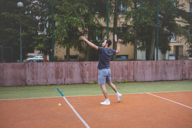 Badminton oyuncusu, açık hava sahasında oynanan bir maçta spiker horozuna vuruyor. Açık alanda kesin teknik ve atletik hareketler sergiliyor. Oyuncu ağaçlar ve kentsel bir zemin ile çevrili..