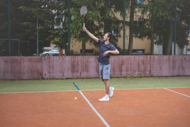 Badminton oyuncusu, açık hava sahasında oynanan bir maçta spiker horozuna vuruyor. Açık alanda kesin teknik ve atletik hareketler sergiliyor. Oyuncu ağaçlar ve kentsel bir zemin ile çevrili..